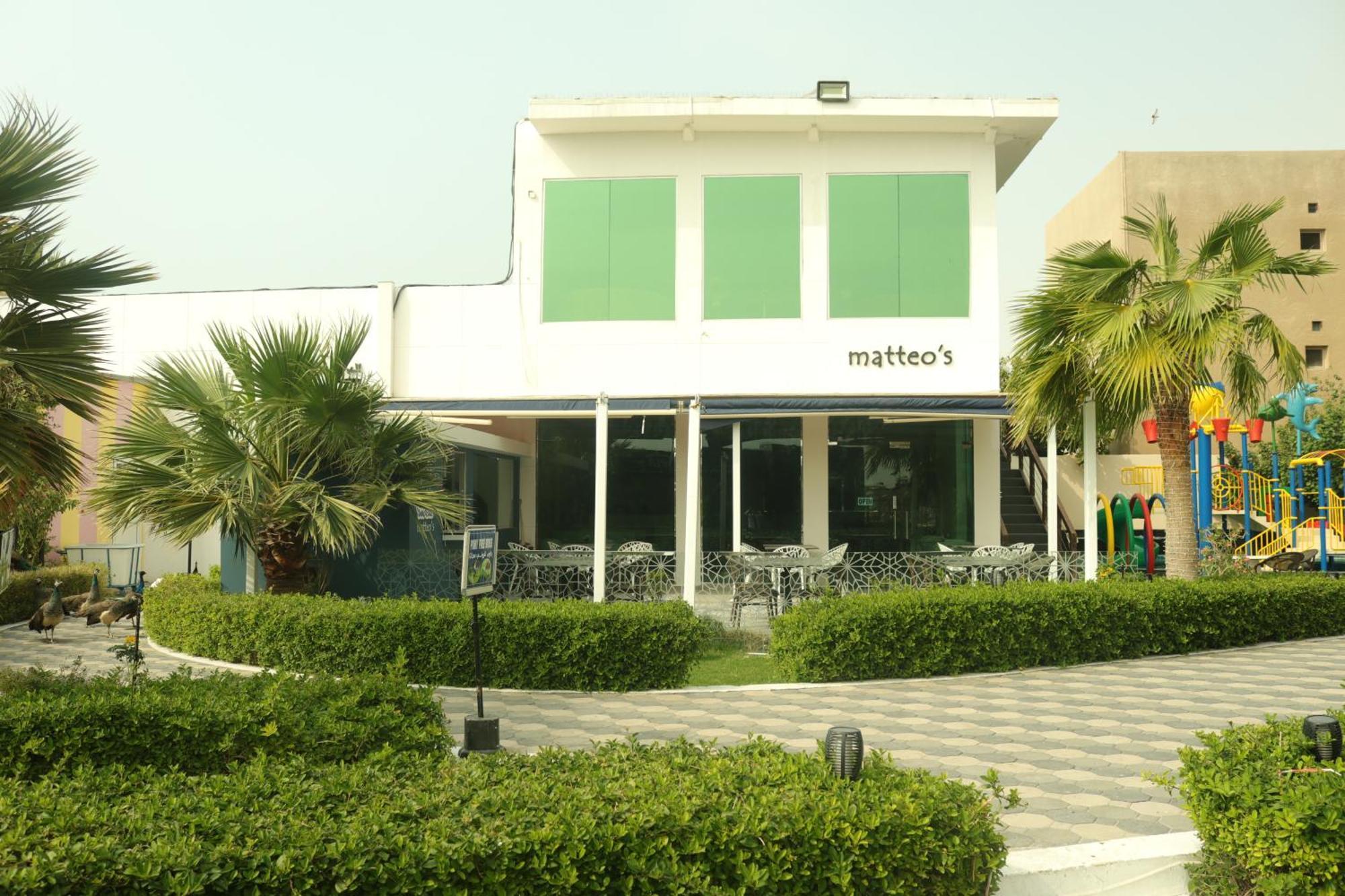 Lagoona Beach Luxury Resort And Spa Manama Exteriör bild The photo shows a building with the name "matteo's" displayed prominently on the front. The structure features large windows and green shutters. There is a landscaped area in front with bushes and palm trees, along with a paved pathway leading up to 