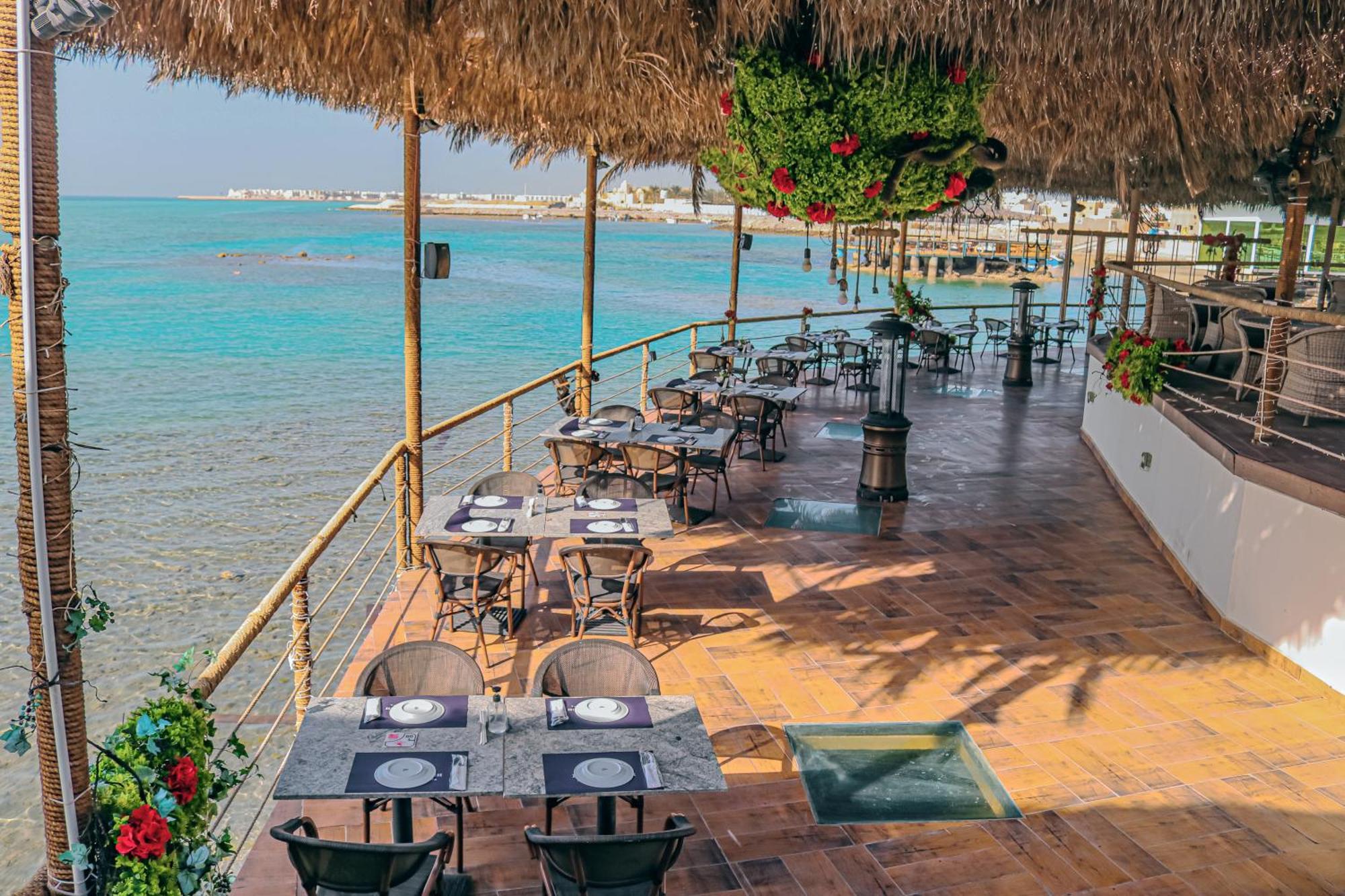 Lagoona Beach Luxury Resort And Spa Manama Exteriör bild The photo shows a seaside restaurant with a spacious outdoor dining area. It features several tables set with plates and utensils, surrounded by lush greenery and decorative plants. The flooring is made of brown tiles, and there are thatched roofs pr