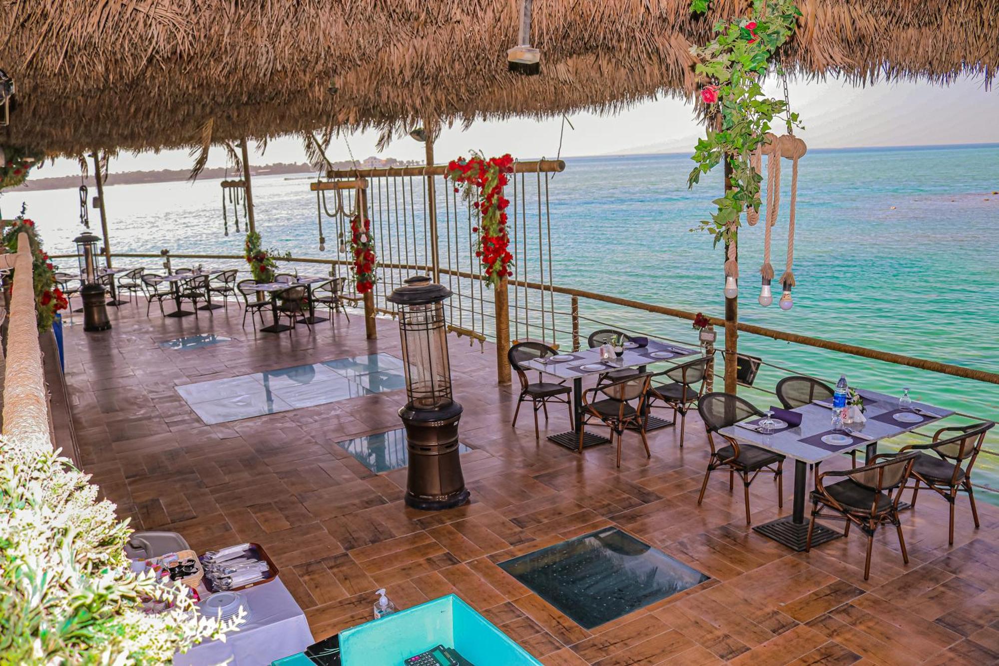 Lagoona Beach Luxury Resort And Spa Manama Exteriör bild The photo depicts a coastal restaurant or dining area with an open-air design. It features a wooden floor and tables set for dining, surrounded by chairs. The space is partially covered by a thatched roof, providing shade while allowing for a view of