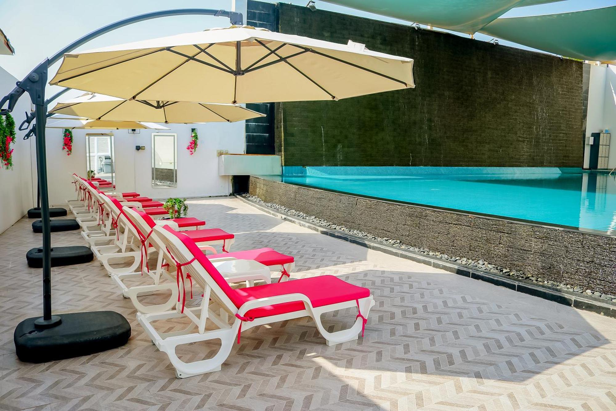 Lagoona Beach Luxury Resort And Spa Manama Exteriör bild The photo shows a sunny poolside area with several lounge chairs arranged in a row. Each chair features red cushions and is positioned under large, beige umbrellas for shade. In the background, there is a beautiful swimming pool with clear blue water