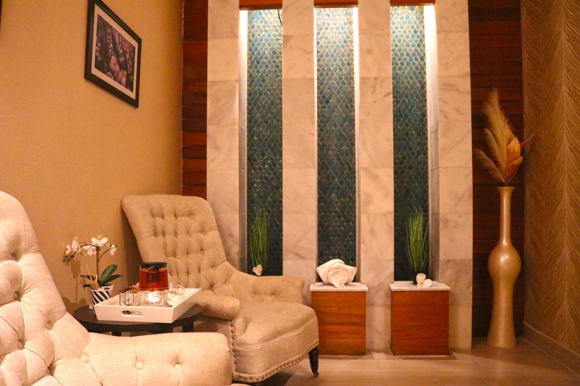 Lagoona Beach Luxury Resort And Spa Manama Exteriör bild The image shows a relaxing interior space, likely a waiting area or lounge. There are two plush, beige armchairs positioned near a small table, which holds a glass pitcher and a cup. The background features tall, elegant windows with decorative blue 