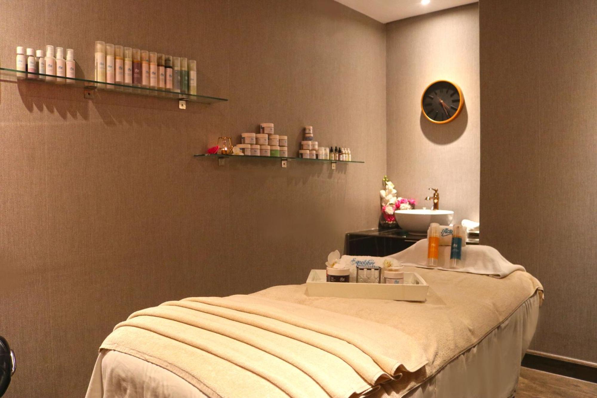 Lagoona Beach Luxury Resort And Spa Manama Exteriör bild The photo shows a spa treatment room. There is a massage table draped with a light-colored blanket, creating a relaxing atmosphere. On the wall, there are shelves displaying various skincare products in bottles and jars. A clock is mounted on the wal
