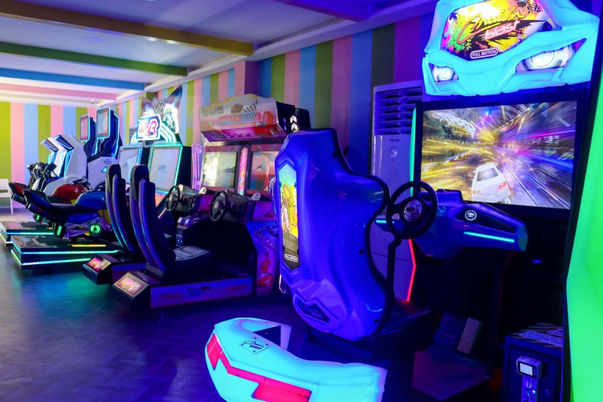 Lagoona Beach Luxury Resort And Spa Manama Exteriör bild The photo shows an arcade area filled with various arcade machines. The setting is illuminated with colorful neon lights, creating a vibrant atmosphere. There are several racing game machines with steering wheels and seats, each accompanied by screen
