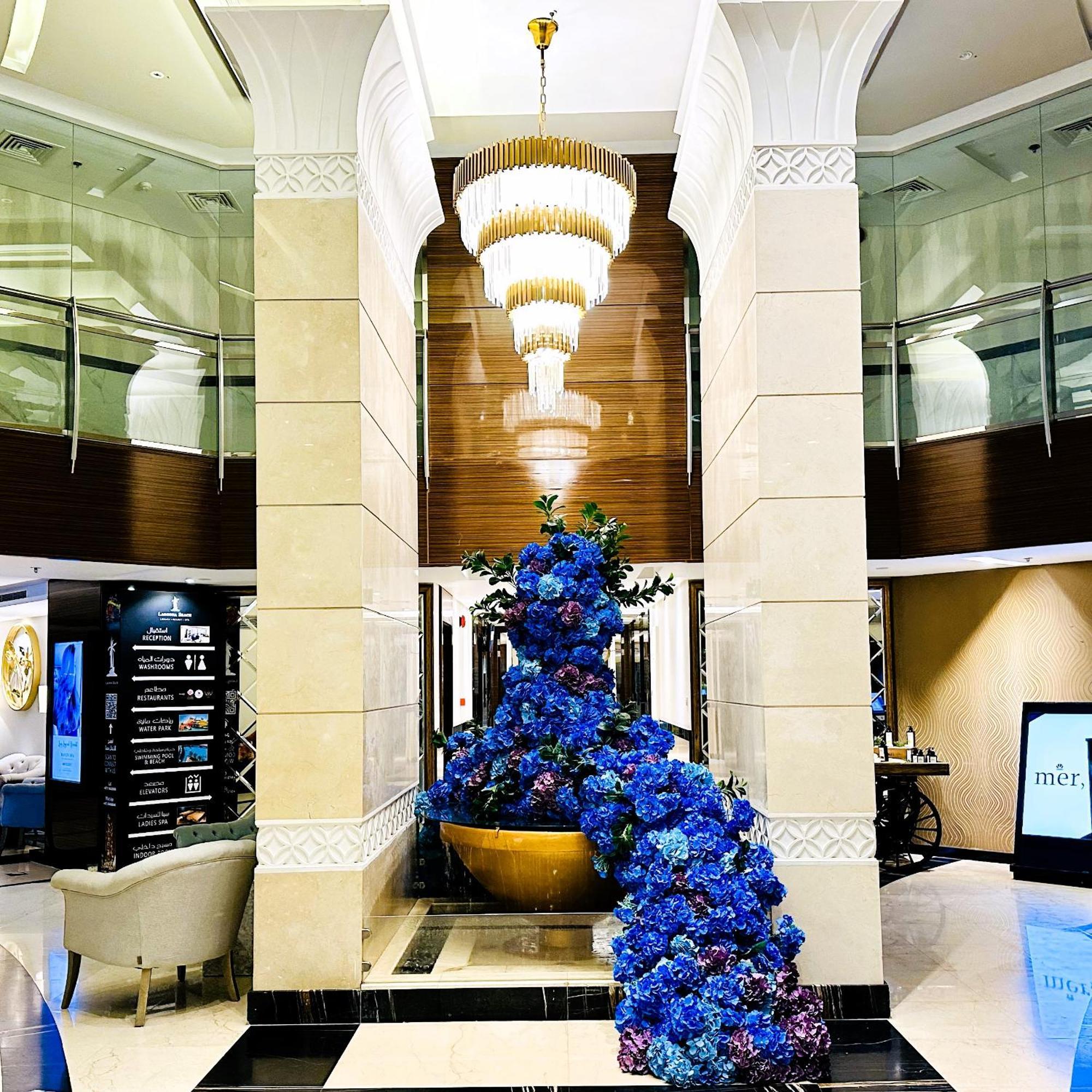 Lagoona Beach Luxury Resort And Spa Manama Exteriör bild The photo depicts an elegant lobby or entrance area of a building. Central to the scene is a large floral arrangement featuring vibrant blue flowers. This arrangement is positioned in a decorative bowl. Above it hangs a stylish chandelier with a laye