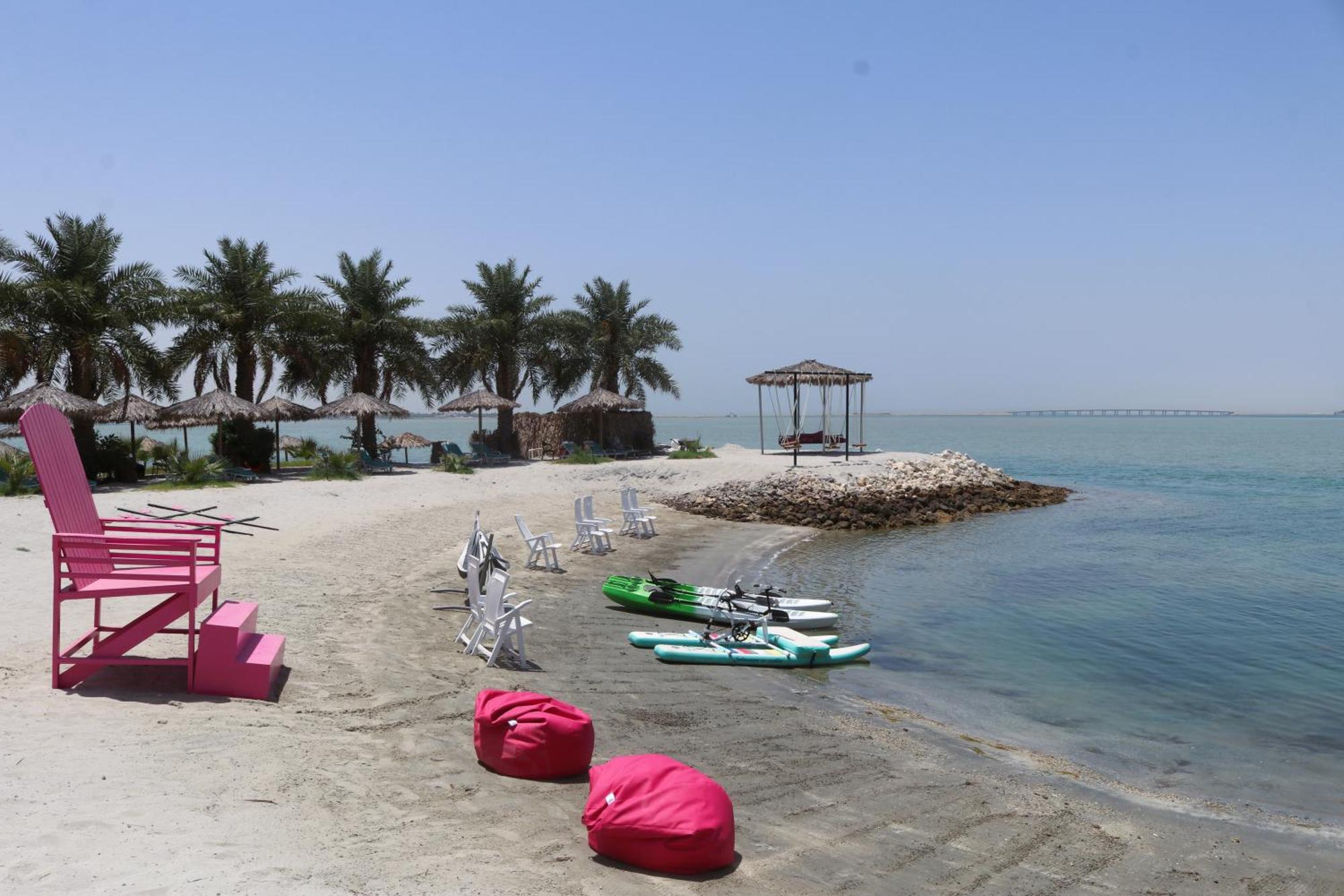 Lagoona Beach Luxury Resort And Spa Manama Exteriör bild The photo depicts a serene beach scene with clear blue water and a sunny sky. In the foreground, there are colorful pink bean bags and a large pink chair on the sand. A few white chairs and loungers are positioned near the water's edge. To the side, 