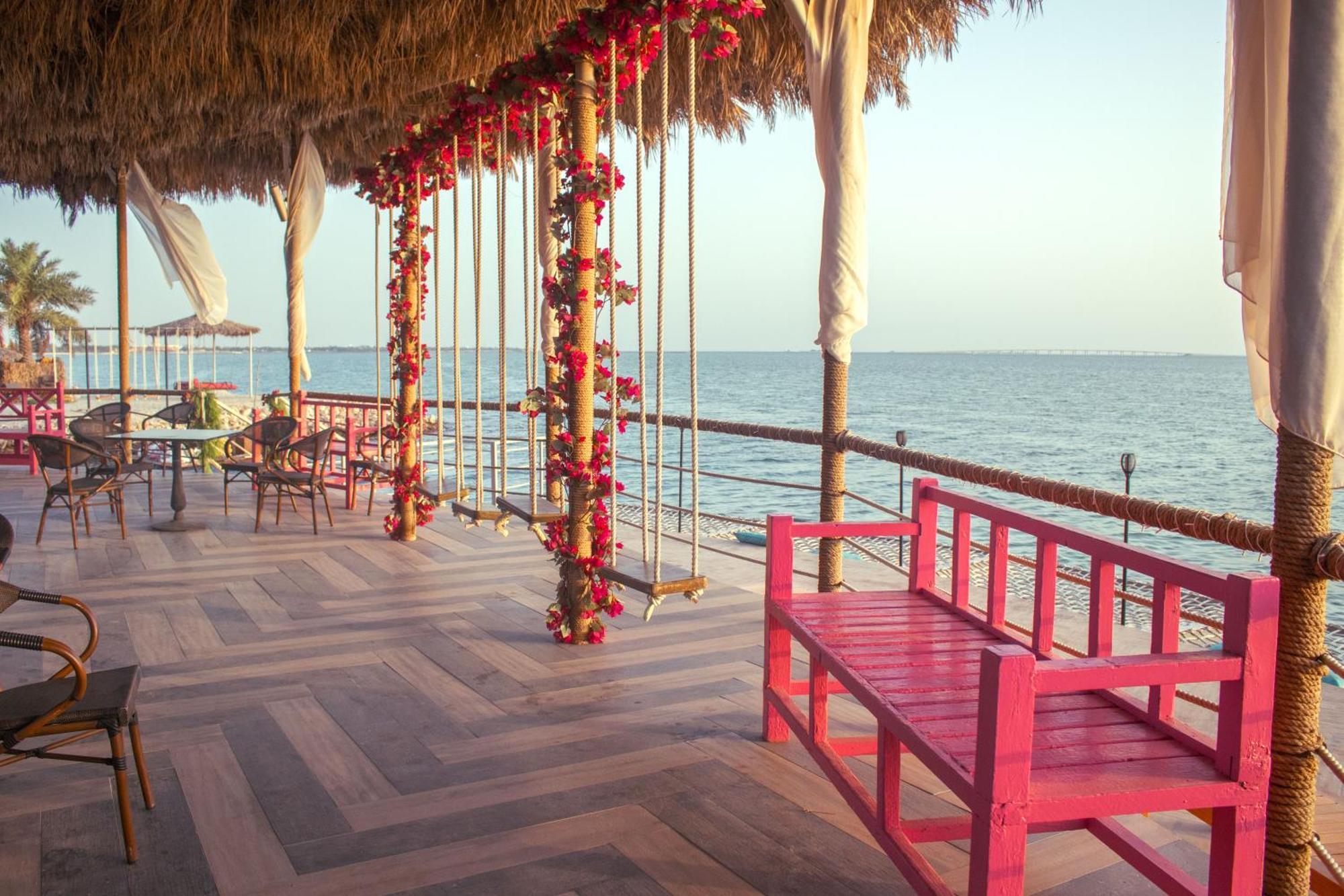 Lagoona Beach Luxury Resort And Spa Manama Exteriör bild The photo depicts a scenic outdoor area by the water, with a wooden deck and a thatched roof. Two swings adorned with flowers are hanging from the ceiling, providing a charming touch to the space. In the foreground, there is a bright pink bench, addi