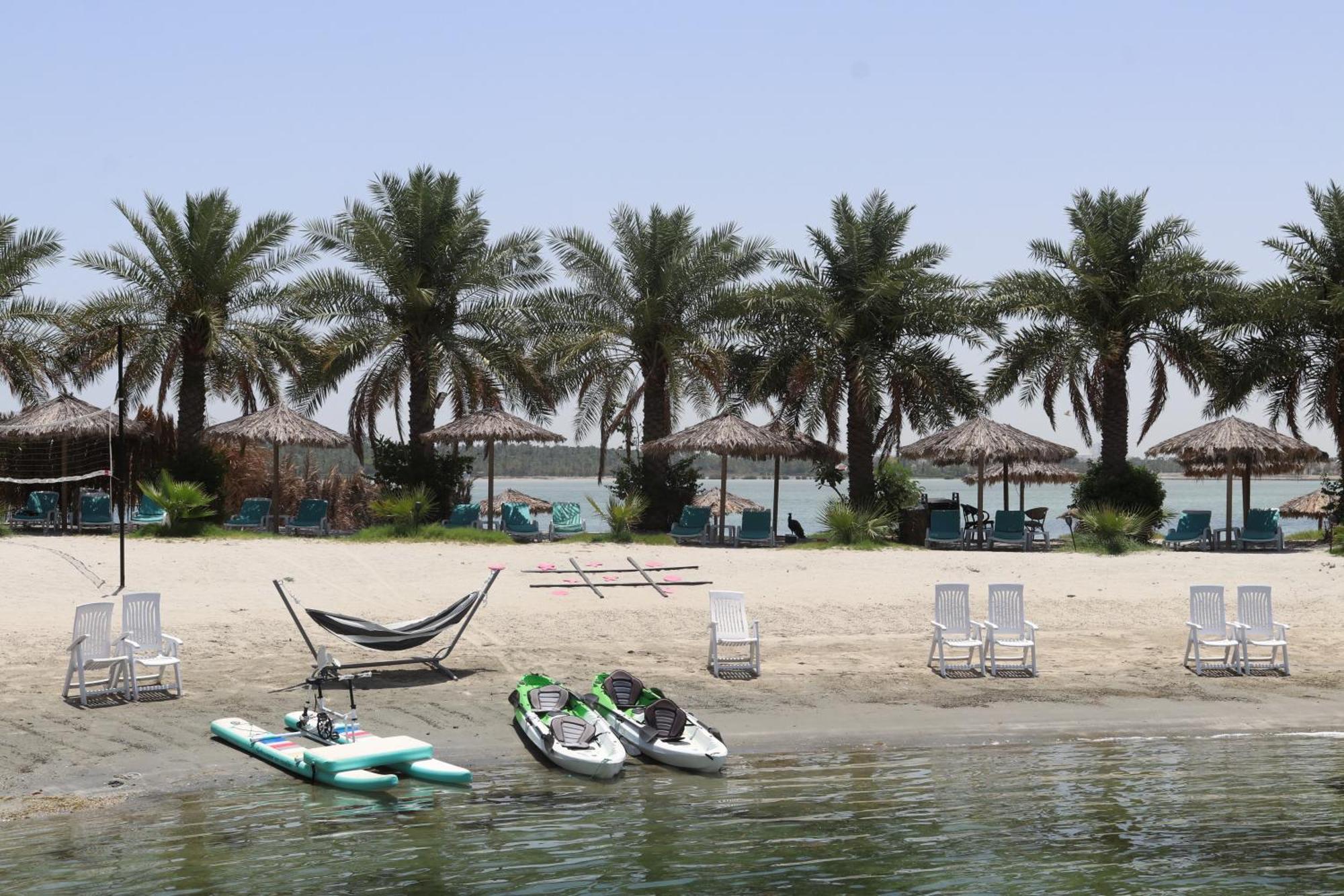 Lagoona Beach Luxury Resort And Spa Manama Exteriör bild The photo depicts a serene beach scene with several palm trees lining the shore. There are thatched umbrellas providing shade, and a few lounge chairs are positioned on the sand. A hammock is set up between two supports, inviting relaxation. In the f