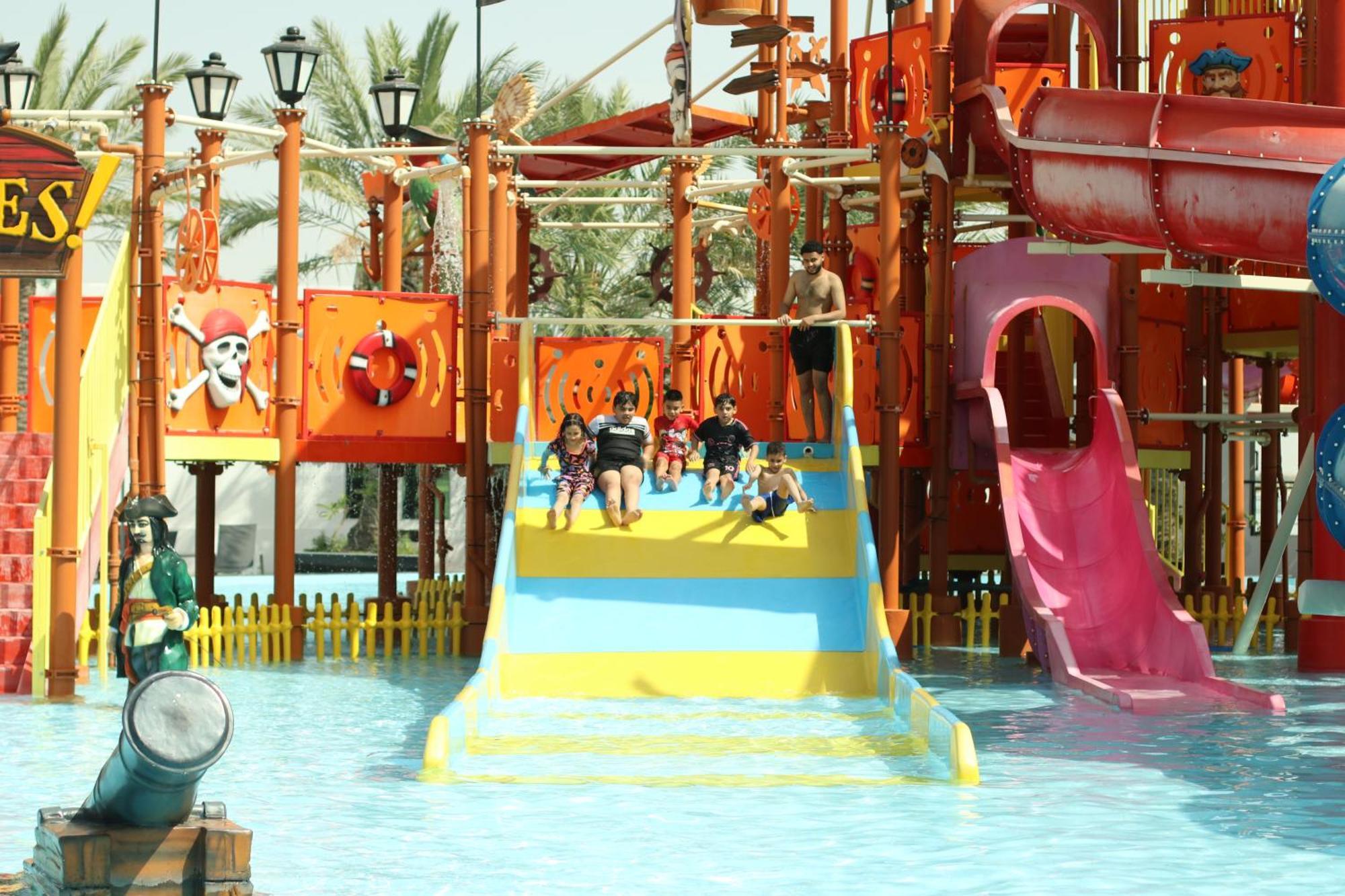 Lagoona Beach Luxury Resort And Spa Manama Exteriör bild The photo shows a colorful water park scene featuring a play area with multiple slides. There are children enjoying themselves, sliding down bright yellow and pink slides into a pool of water. The structure has pirate-themed decorations, including a 