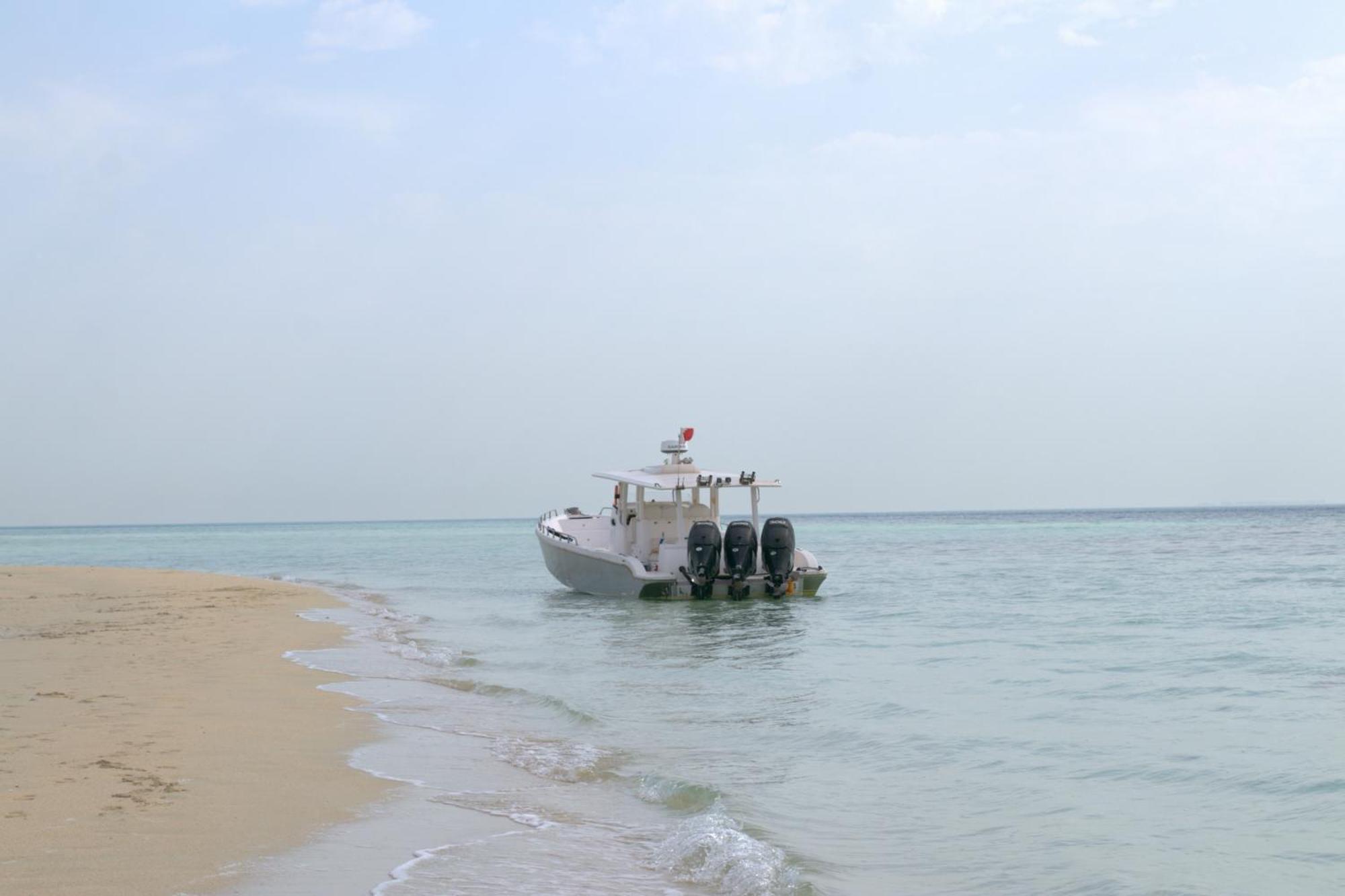 Lagoona Beach Luxury Resort And Spa Manama Exteriör bild The photo depicts a small boat anchored near a sandy beach, partially submerged in calm, clear water. The boat is white and features three outboard motors on its stern. In the background, the sea stretches out toward the horizon under a light, cloudy