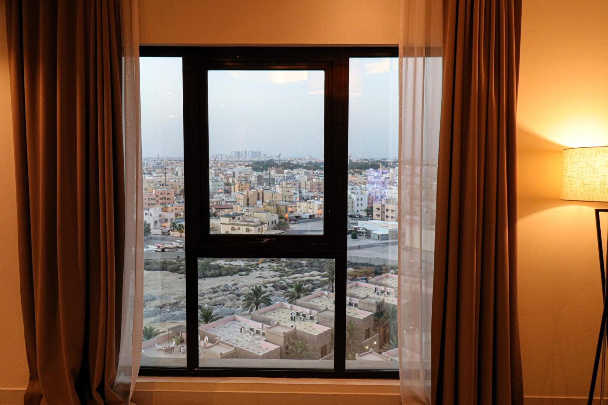 Lagoona Beach Luxury Resort And Spa Manama Exteriör bild The photo shows a window with a black frame and sheer curtains. Through the window, there is a view of a cityscape, displaying a densely populated area with various buildings and rooftops. The sky appears slightly overcast, suggesting a muted lightin