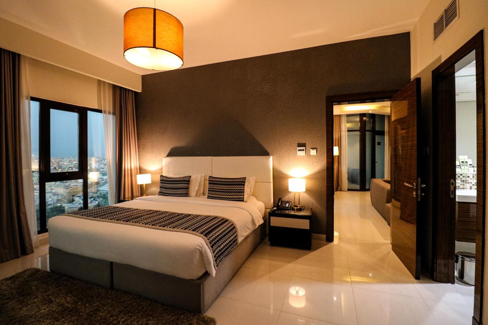 Lagoona Beach Luxury Resort And Spa Manama Exteriör bild The photo depicts a modern hotel room or suite. It features a large bed with a white and gray color scheme, accented with decorative pillows. On either side of the bed, there are sleek bedside lamps providing warm lighting. The walls are a dark color