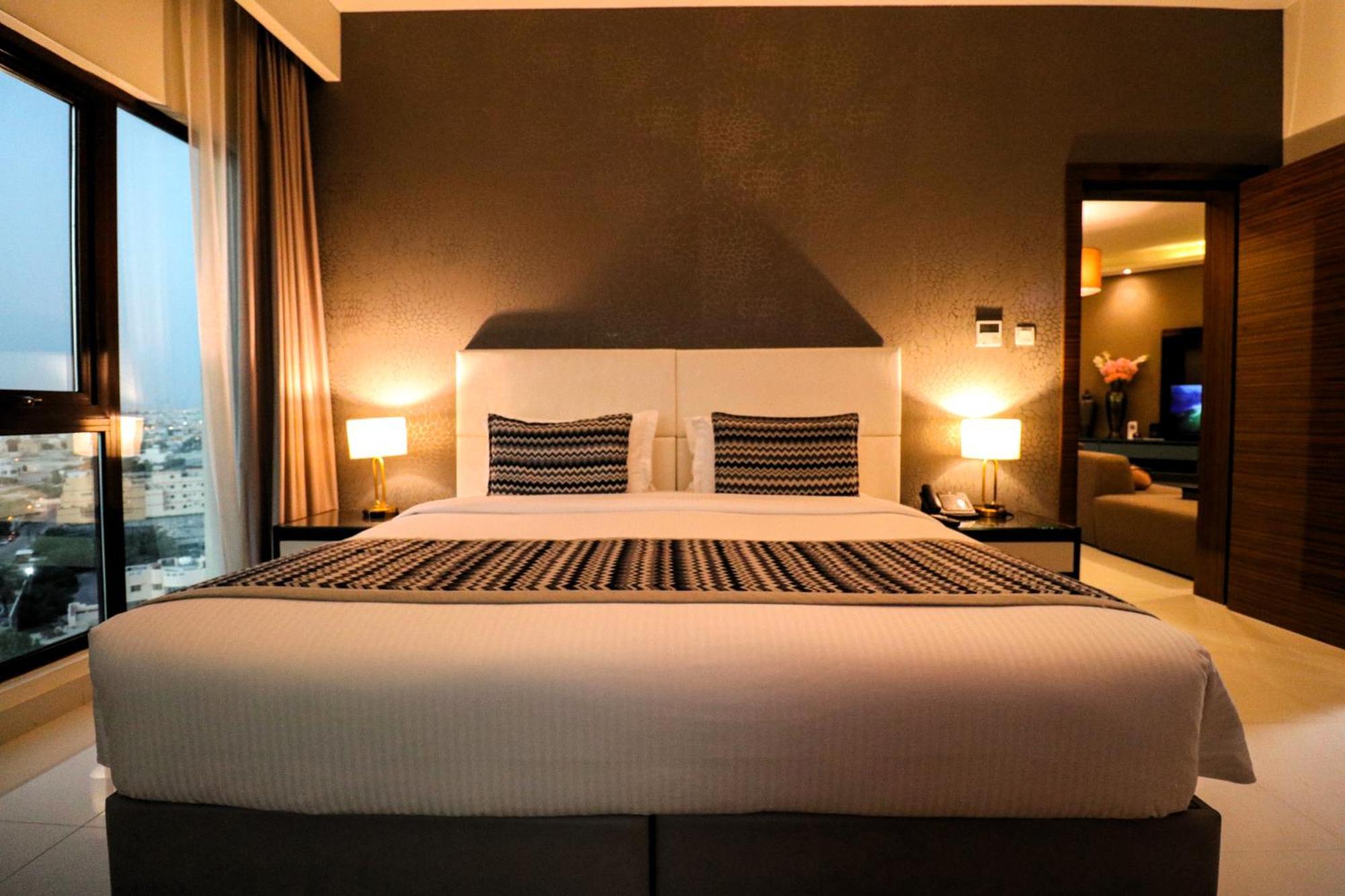 Lagoona Beach Luxury Resort And Spa Manama Exteriör bild The photo depicts a modern hotel room featuring a large bed with a white comforter and two decorative pillows. The headboard is light-colored, and the walls are painted a dark hue. On either side of the bed, there are bedside tables with lamps. The r