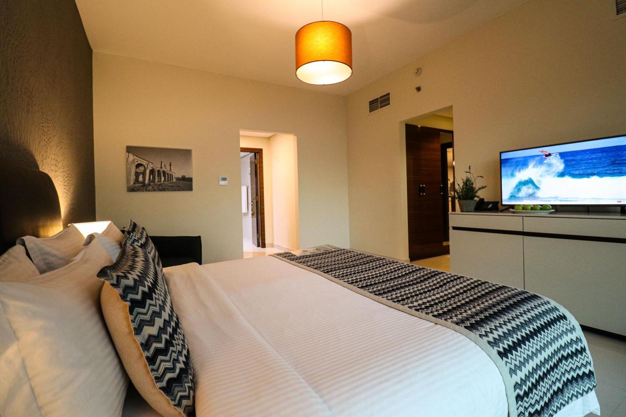 Lagoona Beach Luxury Resort And Spa Manama Exteriör bild The photo shows a well-decorated hotel room. There is a large bed with white linens and decorative pillows featuring a zigzag pattern. On the wall beside the bed is a piece of artwork, and there is a modern television on a white furniture unit opposi