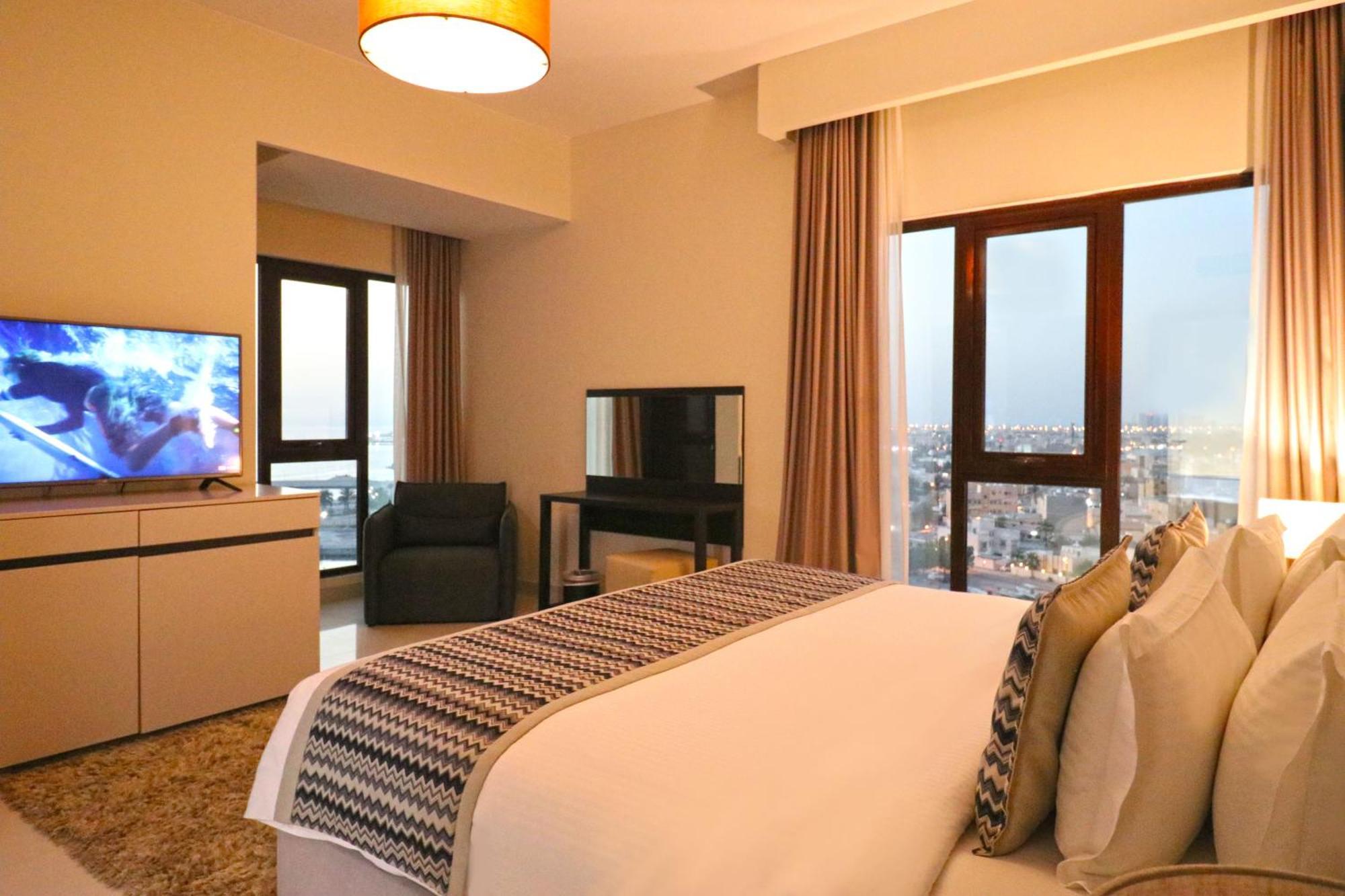 Lagoona Beach Luxury Resort And Spa Manama Exteriör bild The photo shows a modern hotel room or suite. It features a neatly made bed with decorative pillows in the foreground. To the left, there is a TV on a cabinet, and further back, a comfortable chair is situated in front of a large window that likely o