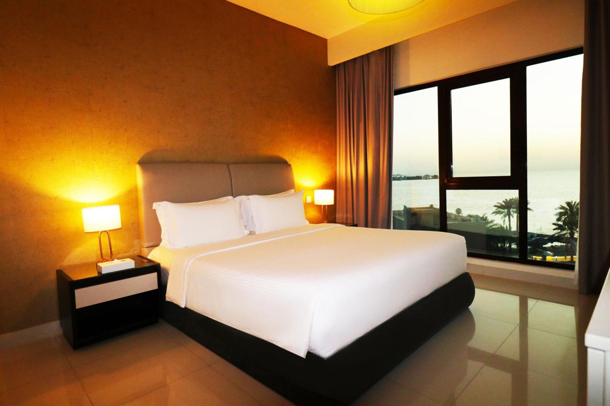 Lagoona Beach Luxury Resort And Spa Manama Exteriör bild The photo shows a modern hotel bedroom with a large, comfortable bed dressed in white linens. The room features warm lighting, including bedside lamps, creating a cozy atmosphere. There are large windows with a view of the outdoors, possibly overlook