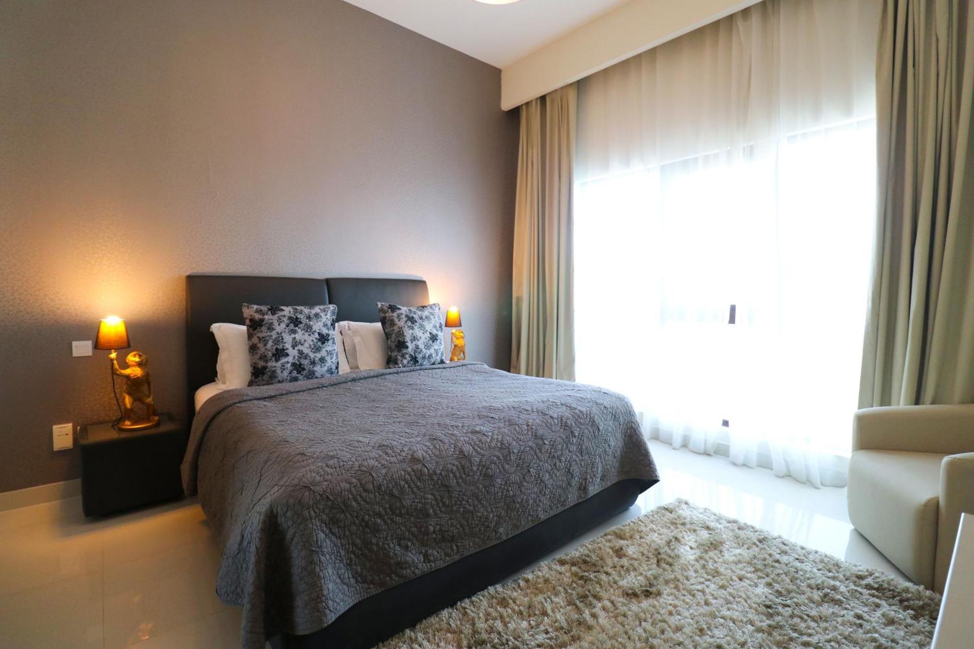 Lagoona Beach Luxury Resort And Spa Manama Exteriör bild The photo shows a stylish bedroom with a modern design. It features a large bed with a dark gray bedspread and decorative pillows. On either side of the bed are two wooden nightstands, each with a lamp. There is a large window covered with light, flo